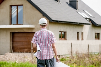 Man with hard hat and architect drawings walking towards half built house