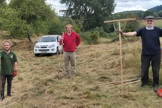 Three men stood apart facing camera holding wooden rakes in hay field