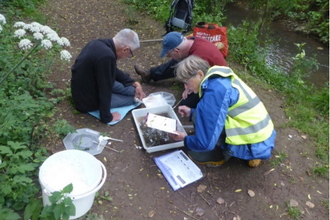 Three people sat beside a stream on a path with a white tray, bucket and paperwork