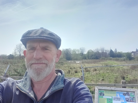 Man wearing a cap looking at camera  with notice board and wild area of grass, water and trees behind