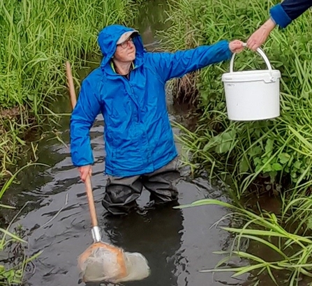 Woman in blue coat stood in stream passing up a white bucket 