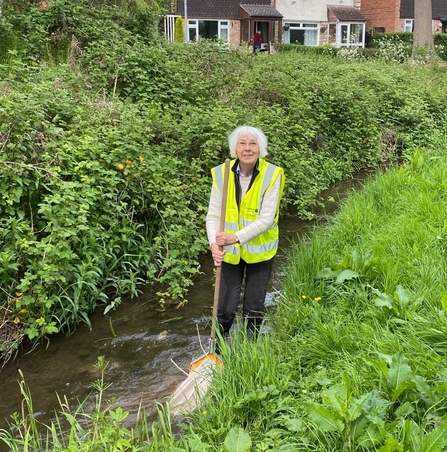 Woman in a hi-vis tabard stood in a stream with houses in the background