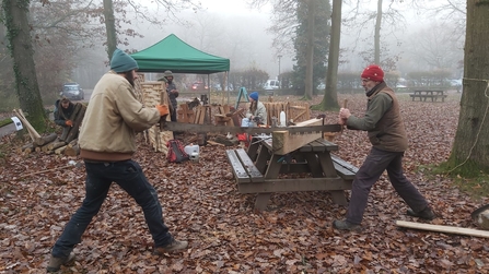Two men using a long saw to cut a plank of wood laid on a picnic bench