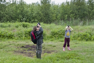 Two children in a grassy outdoor area looking up with binoculars