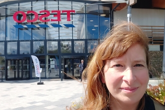 Headshot of woman with red hair stood in front of large building with TESCO logo across the front