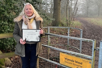 Woman with blond hair wearing a khaki coat stood by gate at entrance to wood holding certificate