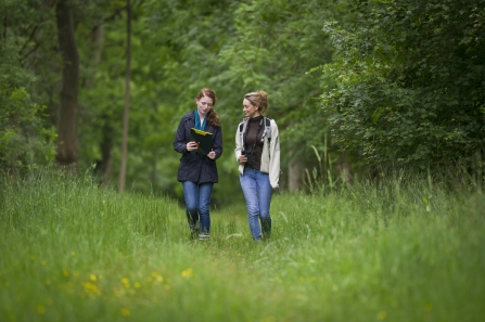 Two women walking together along a grassy path with trees behind