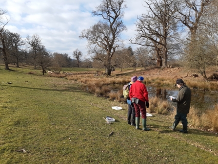 Three people stood by pond in grassland dotted with trees; winter