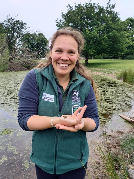 Woman stood in front of pond, smiling at camera cradling a tiny toad in one hand