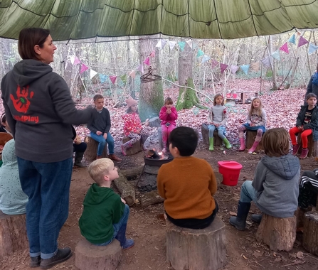 Group of children sitting around a fire on tree stumps under a canopy 