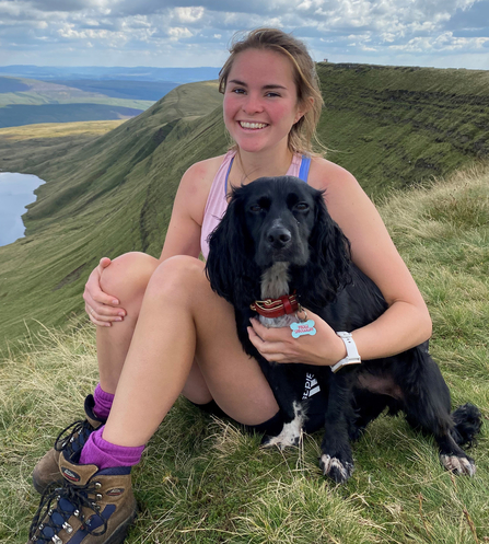 Woman sat with black dog on top of a mountain with green hills and lake visible beyond, white clouds in a blue sky above.