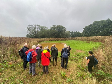 Group of around a dozen people stood by a pond with vegetation surrounding