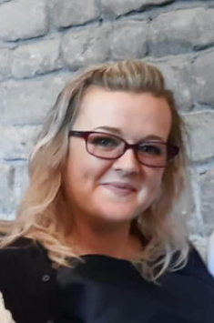 Head and shoulders portrait of a woman with blond hair and red spectacles smiling at the camera