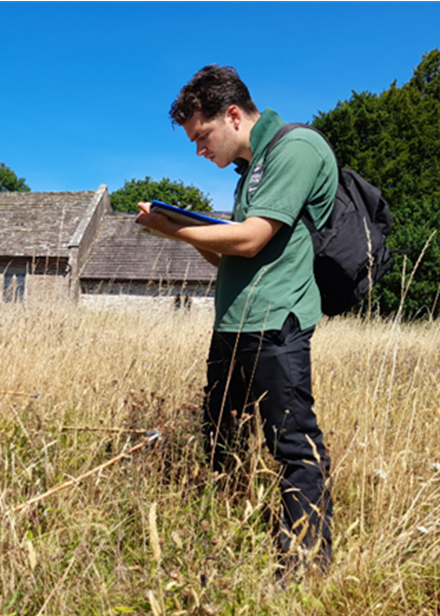 Man with dark hair wearing a green t-shirt holding a clipboard stood in long grass with stone building and trees behind