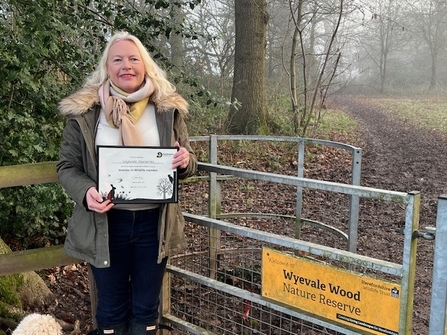 Woman with blond hair wearing a khaki coat stood by gate at entrance to wood holding certificate